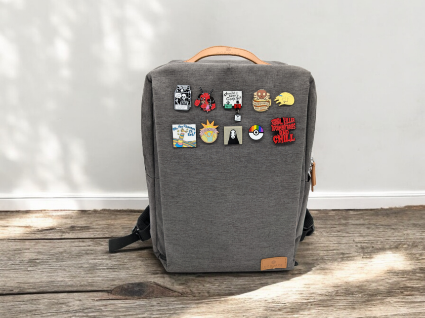 A grey backpack covered in brightly coloured pin badges sits on a wooden floor against a white wall backdrop. The sun creates a warm feeling around the bag.